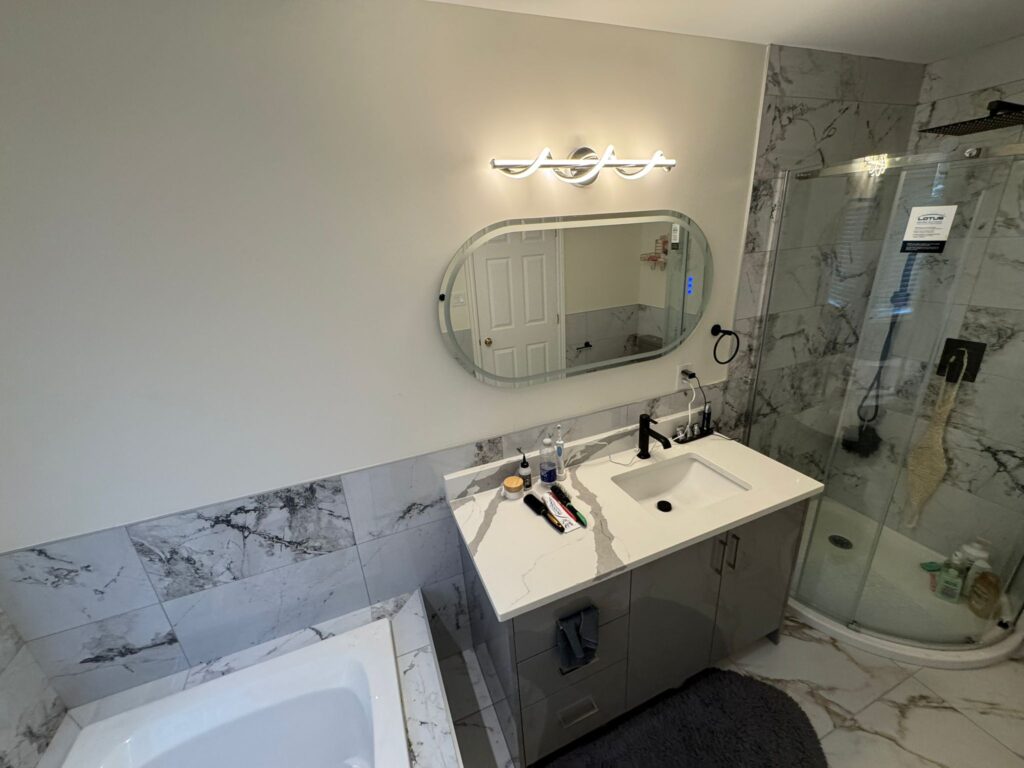 Modern bathroom remodel with grey vanity, white quartz countertop, oval LED mirror, corner glass shower, and white marble-look porcelain tiles covering floor and walls.