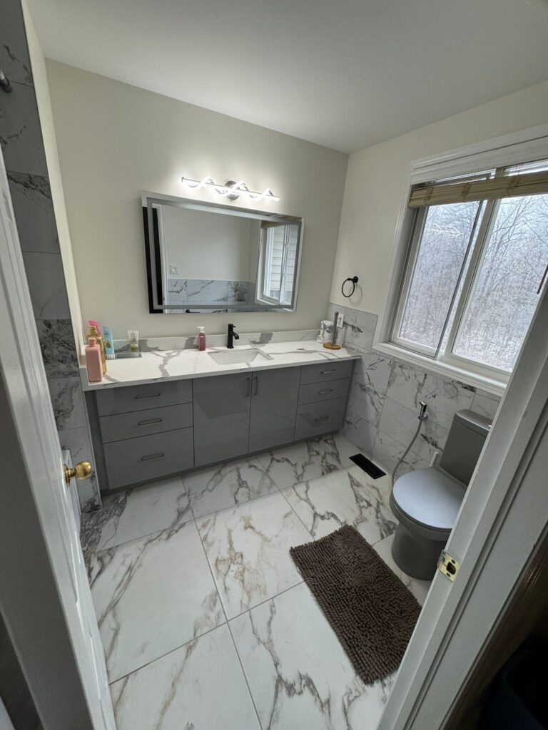 Modern bathroom renovation with marble-look porcelain tiles, grey vanity with quartz countertop, corner glass shower, LED mirror, and unique grey toilet.