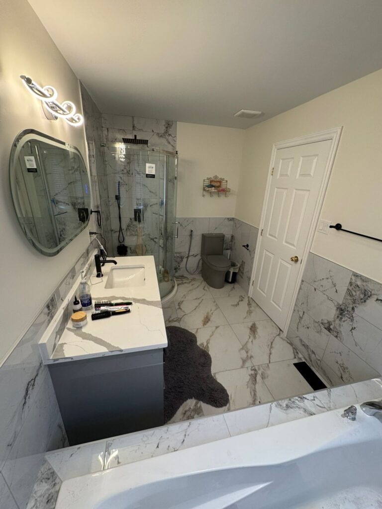 Modern bathroom renovation with marble-look porcelain tiles, grey vanity with quartz countertop, corner glass shower, LED mirror, and soaking tub.