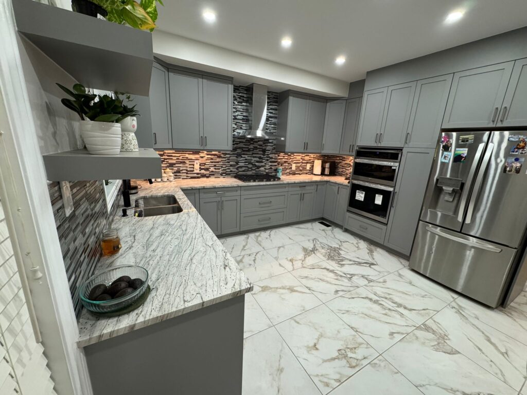 Modern grey kitchen renovation featuring stainless steel double wall oven, floating shelves with decor, linear mosaic backsplash, and marble-look floor tiles.