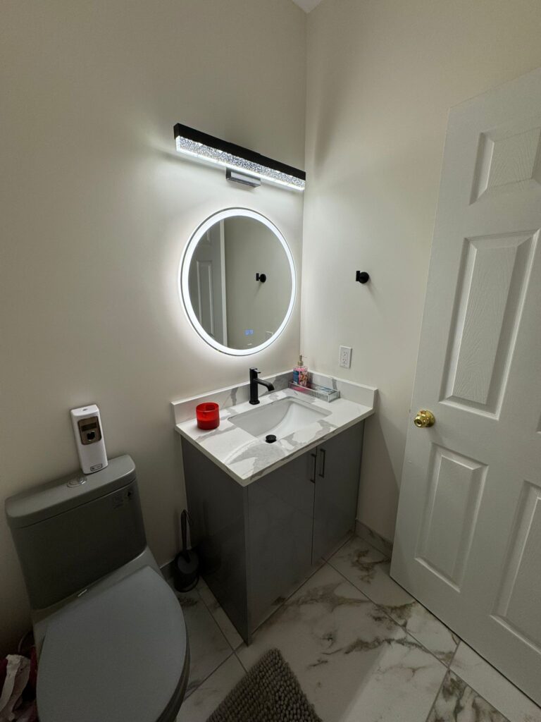 Modern powder room renovation featuring a small grey vanity, circular LED mirror, unique grey toilet, crystal vanity light, and marble-look porcelain flooring.